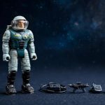 A vintage astronaut figure stands beside miniature accessories on a dark surface.